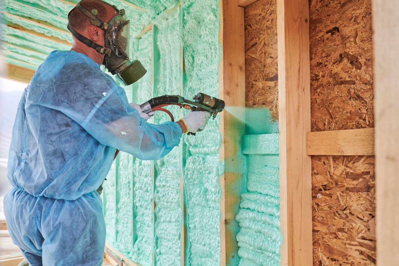 Male builder insulating wooden frame house. Man worker spraying polyurethane foam inside of future cottage, using plural component gun. Construction and insulation concept.